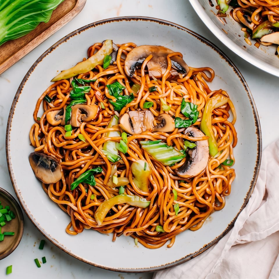 Stir-fried noodles with mushrooms and bok choy served on a plate, garnished with scallions and surrounded by chopsticks.