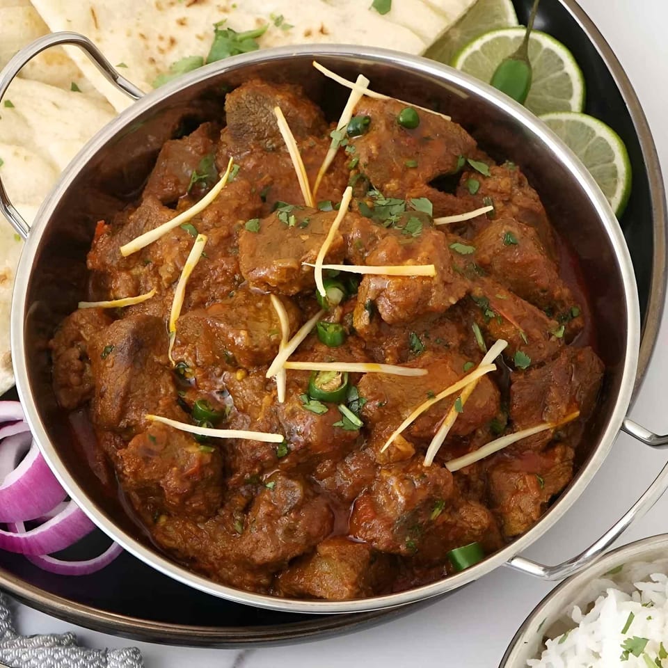 Spiced meat curry garnished with ginger, green chilies, and cilantro, served with naan, lime wedges, onions, and rice.