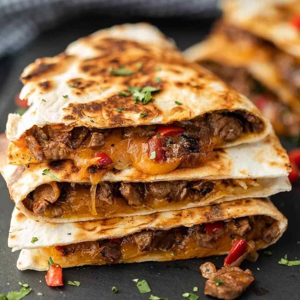 Stack of grilled beef quesadillas filled with melted cheese, peppers, onions, and garnished with cilantro.